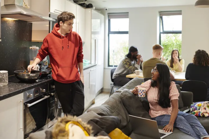 A group of students are gathered in a shared common space. 2 students are chatting while they cook.